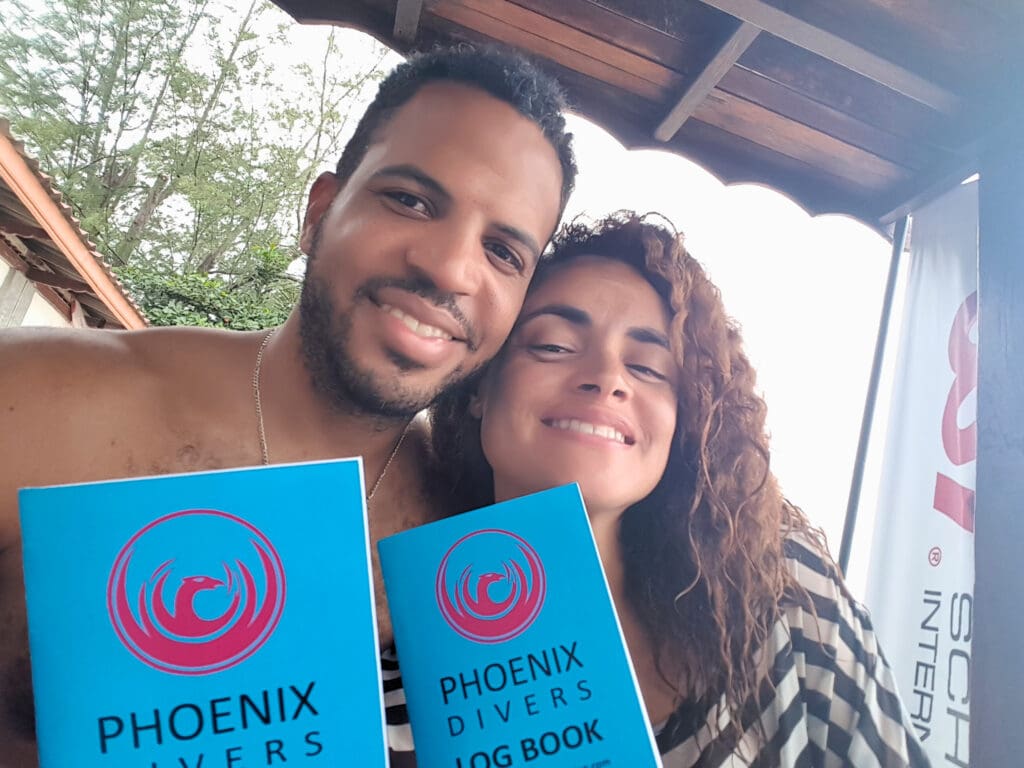 a couple smile into the camera with their dive books on display