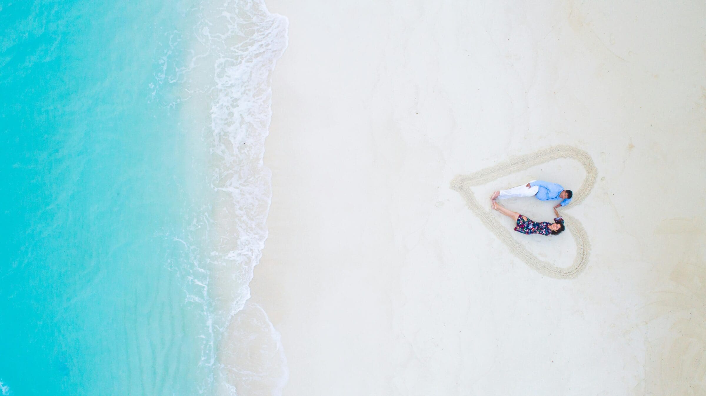 Aerial view of beach with hear drawn in sand and a couple holding hands