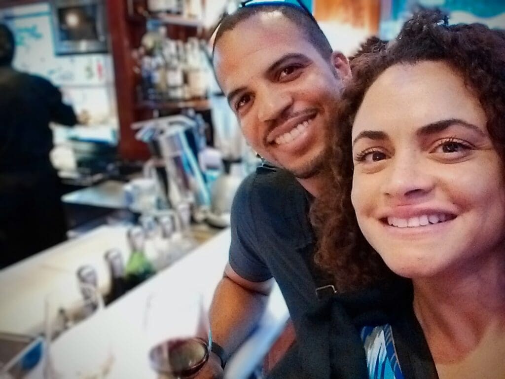 man and woman smile into the camera with 2 glasses of wine on the bar.