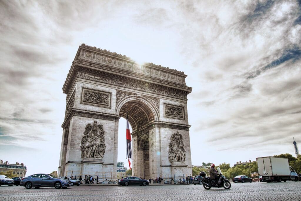 A stunning daytime view of the Arc de Triomphe in Paris with traffic and the Eiffel Tower in the background.
