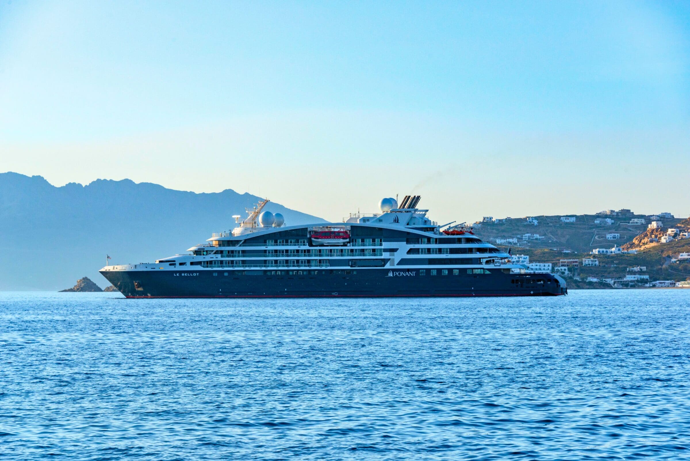 Majestic small ship cruise sailing near a coastal town with a scenic mountain backdrop. Mykonos Greece