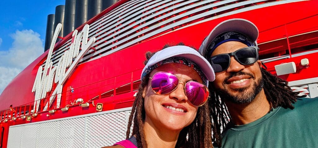 Couple smiling at the camera with Virgin Voyages red lady ship in the background.